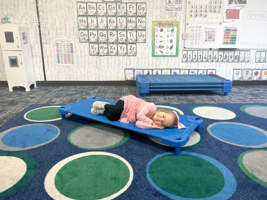 A young child napping comfortably on a blue stackable cot in a classroom setting