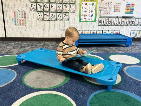 A young child reading on a blue stackable cot in a classroom setting